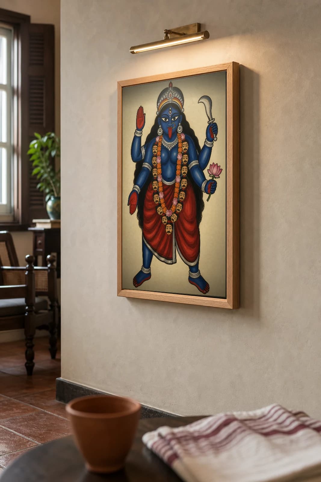 Maa Kali Kalighat Print — the blue goddess in dakshina stance with khadga, lotus and a garland of heads, the namesake deity of Kalighat, devotional Bengal patua brush, hand-numbered collector edition — the framed print shown at a three-quarter angle on a styled gallery wall in a natural-oak frame