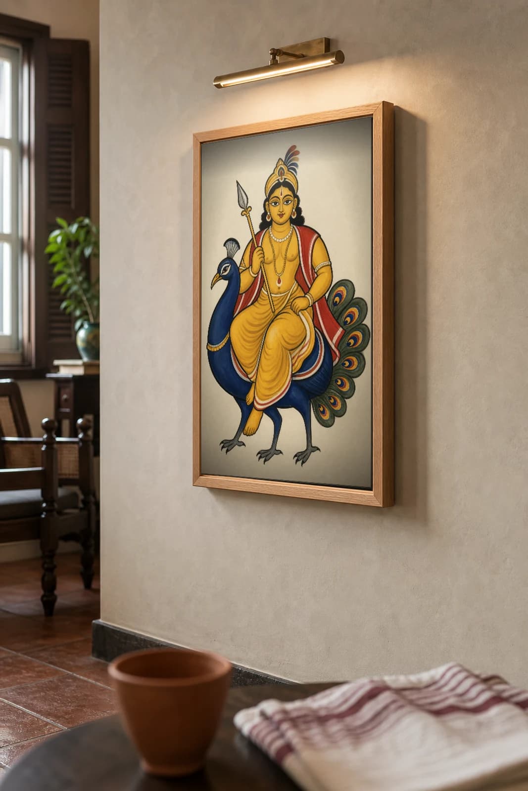 Kartik Kalighat Print — the handsome war-god seated on his peacock with a silver spear, a Bengali household favourite, devotional patua brush, hand-numbered collector edition — the framed print shown at a three-quarter angle on a styled gallery wall in a natural-oak frame