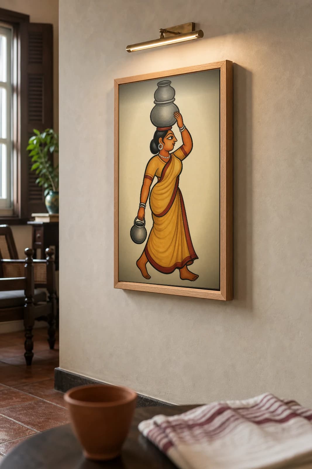 The Milkmaid — Kalighat-style art print of a woman carrying milk pots in a saffron saree, Bengal patua brush · collector edition · framed A2 — the framed print shown at a three-quarter angle on a styled gallery wall in a natural-oak frame
