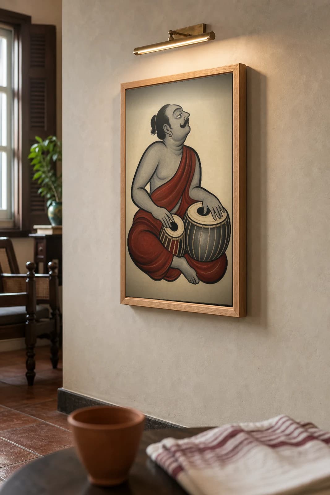 The Ostad — Kalighat-style art print of a seated tabla player mid-bol, Bengal patua brush · collector edition · framed A2 — the framed print shown at a three-quarter angle on a styled gallery wall in a natural-oak frame
