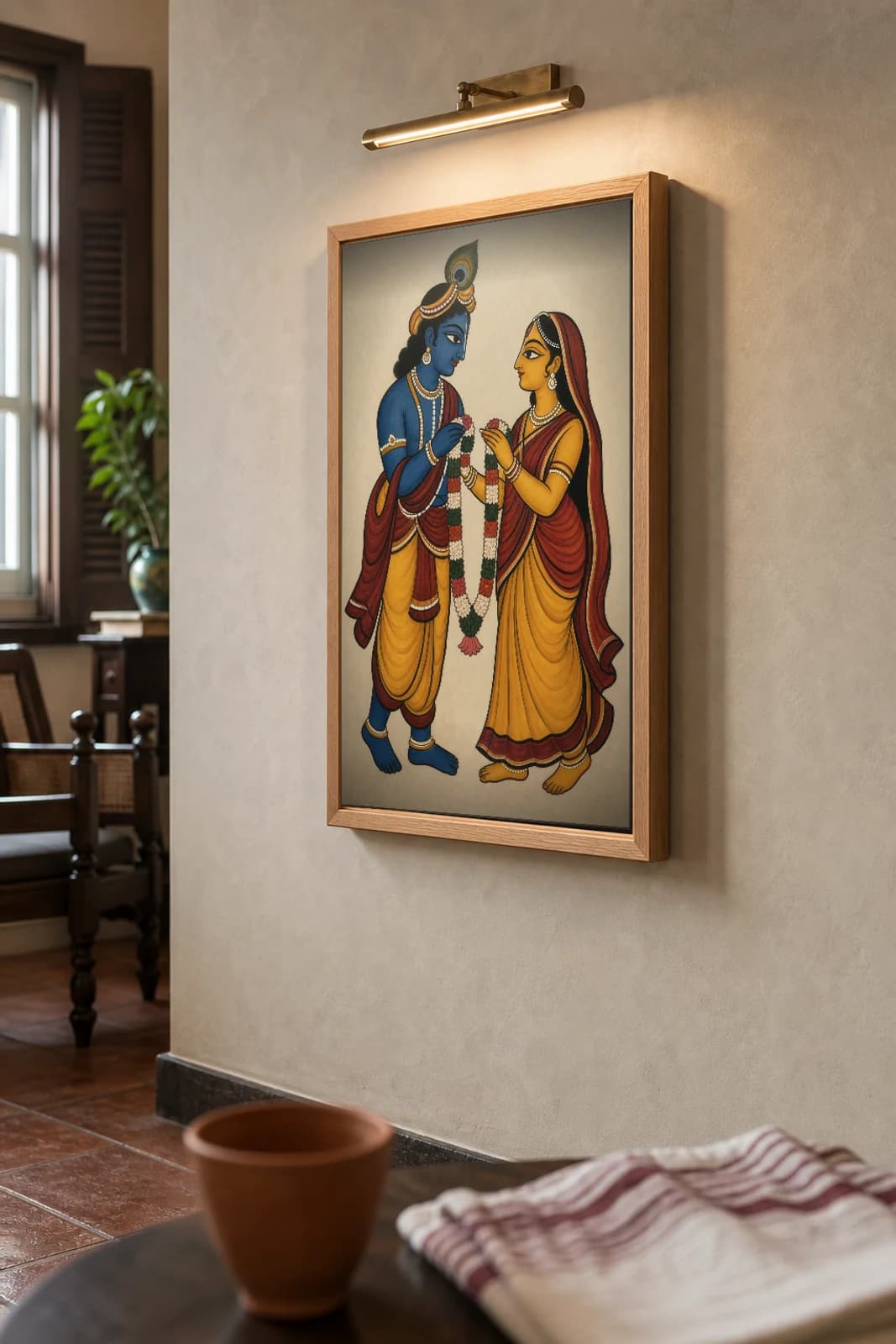 Radha Krishna — Kalighat-style devotional art print of the divine pair exchanging a garland, Bengal patua brush · collector edition · framed A2 — the framed print shown at a three-quarter angle on a styled gallery wall in a natural-oak frame