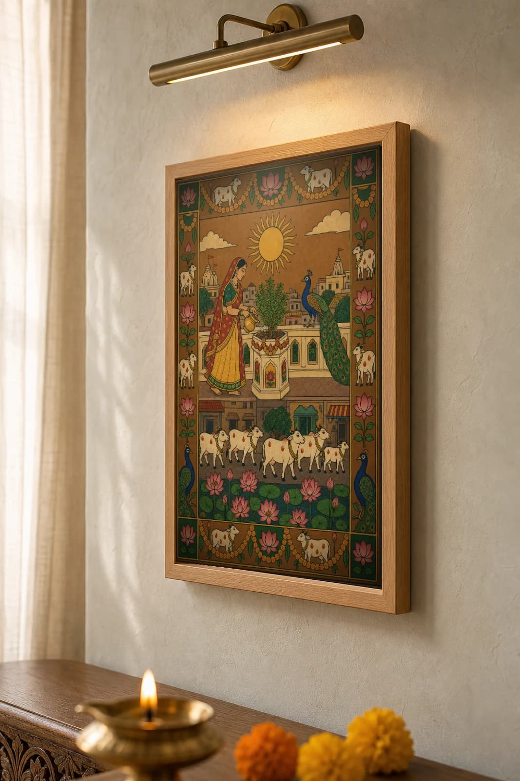 Tulsi Balcony Pichwai-Style Print — a woman watering a tulsi plant on a city terrace under a rising sun, with a peacock and cows in a lotus pond below, on warm ochre cloth — the framed print shown at a three-quarter angle on a styled gallery wall in a natural-oak frame