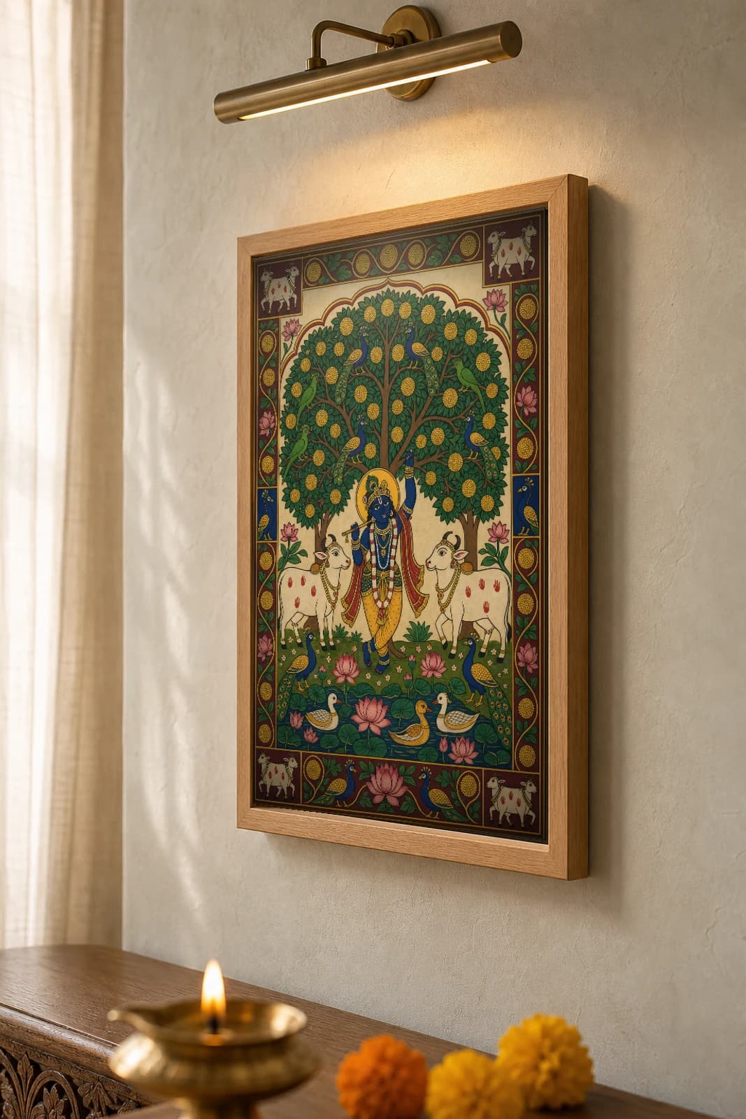 Kadamba Vriksha Pichwai-Style Print — flute Krishna beneath a great yellow-flowering kadamba tree flanked by two cows, with parrots, peacocks and a lotus pond, on warm cream cloth — the framed print shown at a three-quarter angle on a styled gallery wall in a natural-oak frame